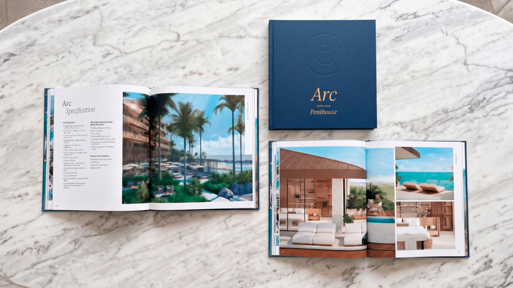 Libro Arc Windward: libro de tapa azul cerrado junto a dos páginas abiertas con fotos del proyecto sobre una mesa de mármol